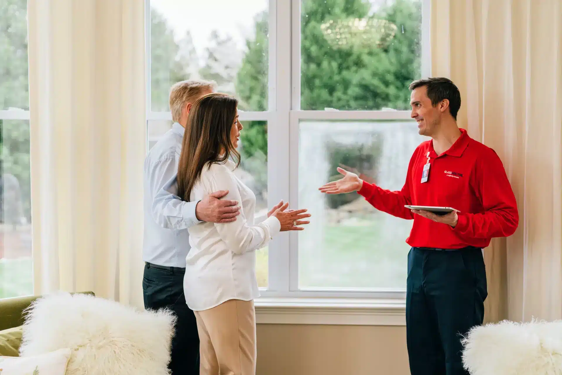 Glass Doctor technician with a tablet consulting with homeowners by a large window during a home glass replacement consultation.