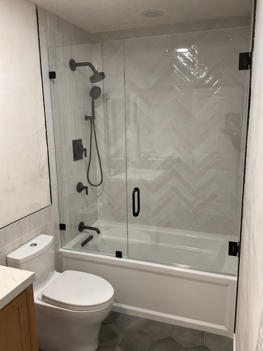 Frameless two-panel glass tub enclosure with matte black hinges and handle installed over a bathtub in a Murrieta bathroom featuring herringbone tile walls.