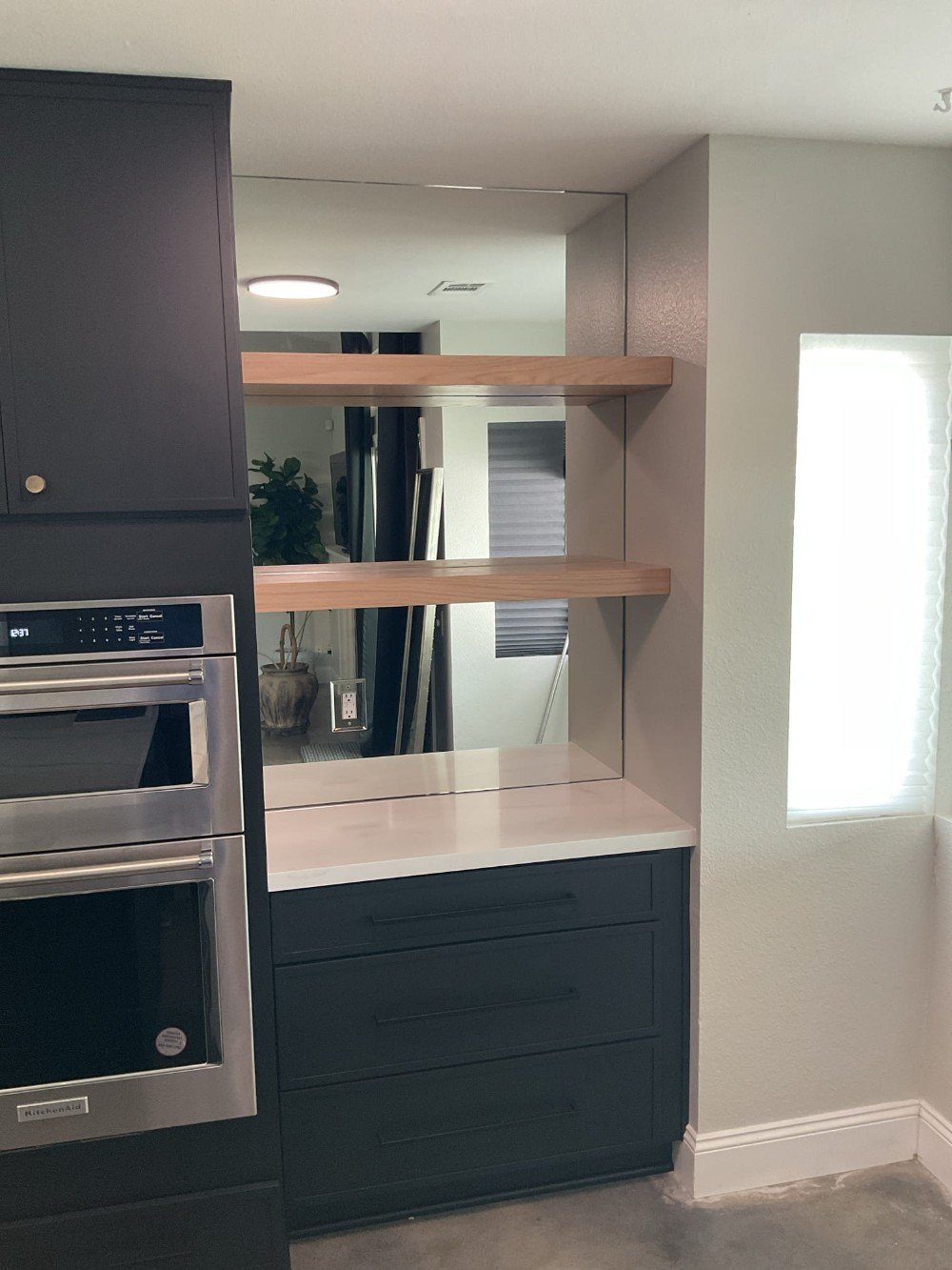 Custom three-tier mirror installation behind floating wooden shelves in a modern kitchen with black cabinetry and stainless steel appliances.