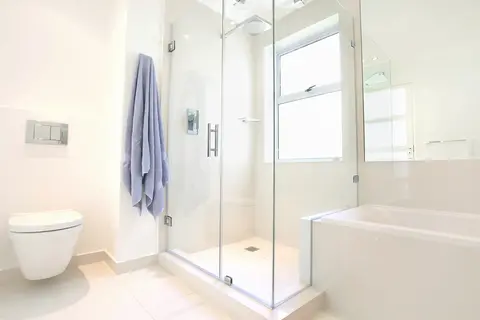 A two sided frameless glass shower enclosure with a swining door in a white bathroom.