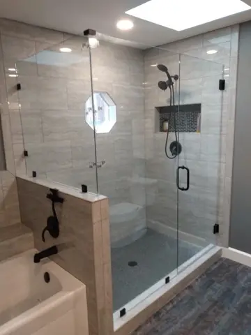 A gray tile shower with frameless glass at 90-degrees, a swinging door, and dark hardware by Glass Doctor of Raleigh