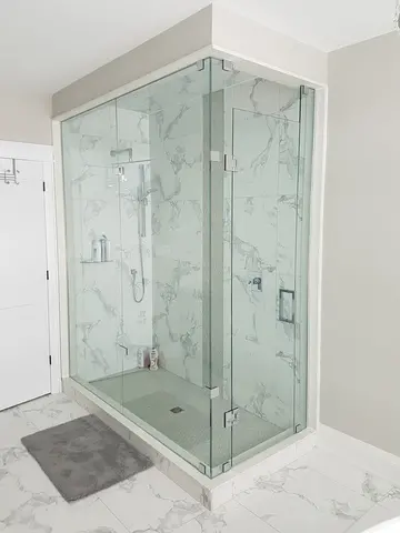 A corner shower with white marble and two sided frameless glass enclosure and a swinging door on one end installed by Glass Doctor of Muskoka.