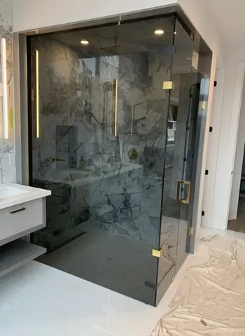 A shower enclosure with 90-degree tinted frameless glass panels and gold hardware by Glass Doctor of Newmarket.
