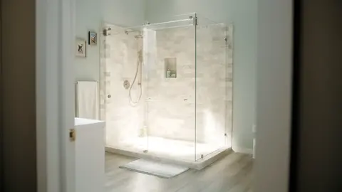 A beige corner shower with a two-sided frameless clear glass enclosure and a swinging door.