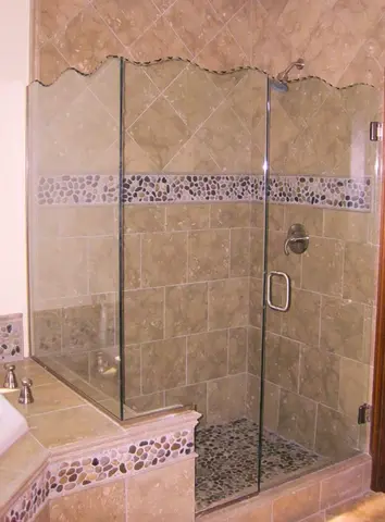 A light brown tiled shower with a two-sided frameless glass enclosure featuring a wavy decorative top edge.