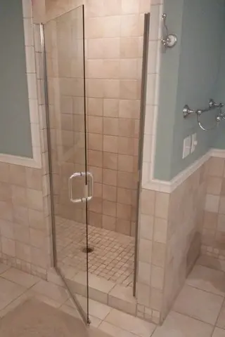 A swinging clear frameless glass door installed on a walk-in shower with beige tiles.