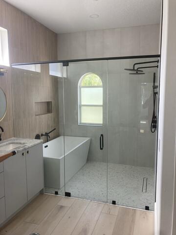 A wet room with a white tub and gray tiles enclosed with clear glass and a black top frame and center door installed by Glass Doctor of Ocala.