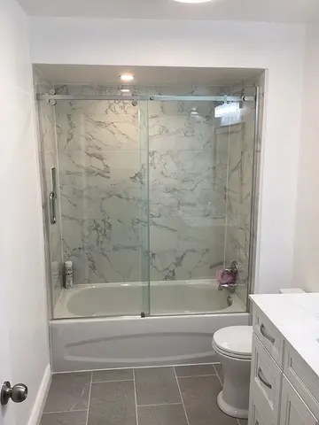 A white bath tub with white and grey marble tiles and a frameless sliding glass enclosure intalled by Glass Doctor of York Region.