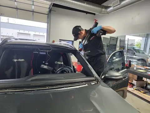 A skilled Glass Doctor Auto of Cedar Park uses an adhesive gun to apply adhesive to a gray car for a windshield replacement.