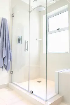 A two sided frameless glass shower enclosure with a swining door in a white bathroom.