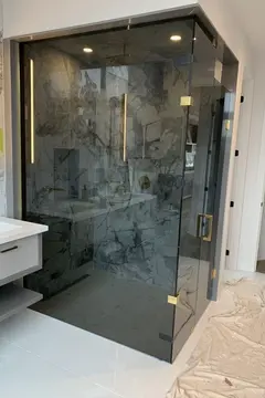 A shower enclosure with 90-degree tinted frameless glass panels and gold hardware by Glass Doctor of Newmarket.