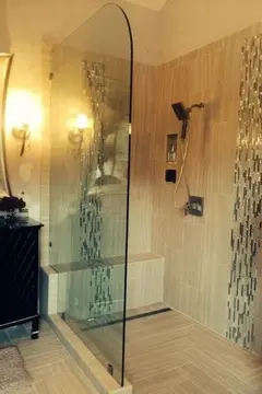 A shower with with brown tiles and a single pane glass panel bath screen with a rounded top corner.