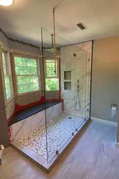 A walk-in shower with white and gray tile and a frameless glass splash panel in front of a bay window from Glass Doctor of Raleigh