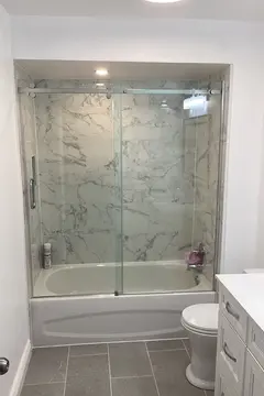 A white bath tub with white and grey marble tiles and a frameless sliding glass enclosure intalled by Glass Doctor of York Region.