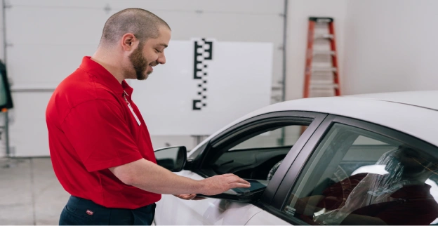 A male Glass Doctor service professional standing beside the driver side window of a white car.