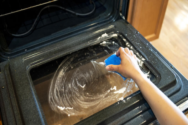 A person using a blue sponge to clean the interior of an oven.