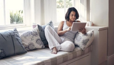 Woman looking at her tablet while lying on a window seat
