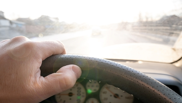 Person driving with a hand on the wheel and looking into the glare from the sun coming through a windshield