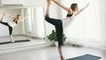Young woman doing stretching exercises on yoga mat in front of a large mirror.